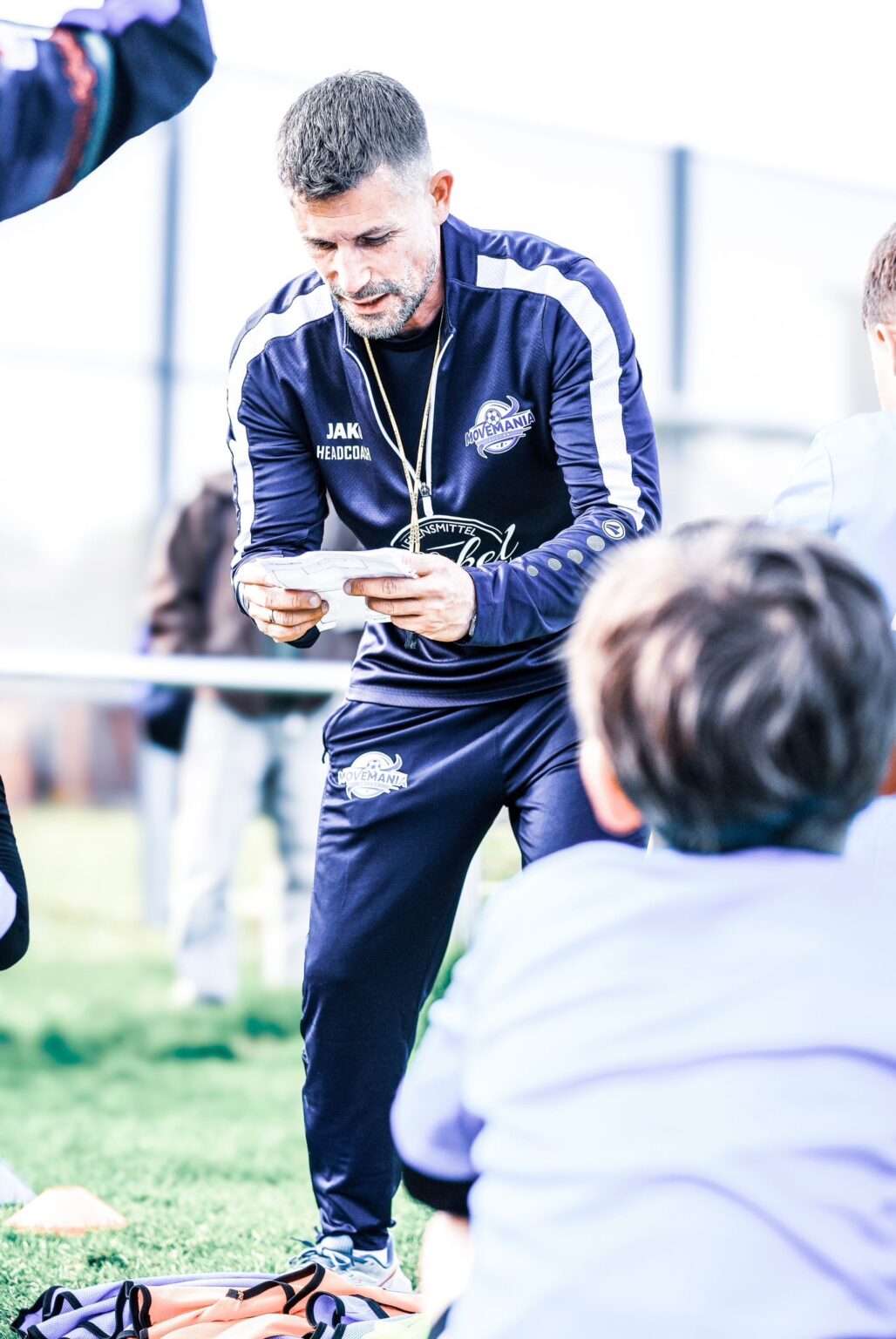 Headcoach in Movemania-Trainingsjacke prüft einen Zettel auf dem Platz, vor ihm ein Kind